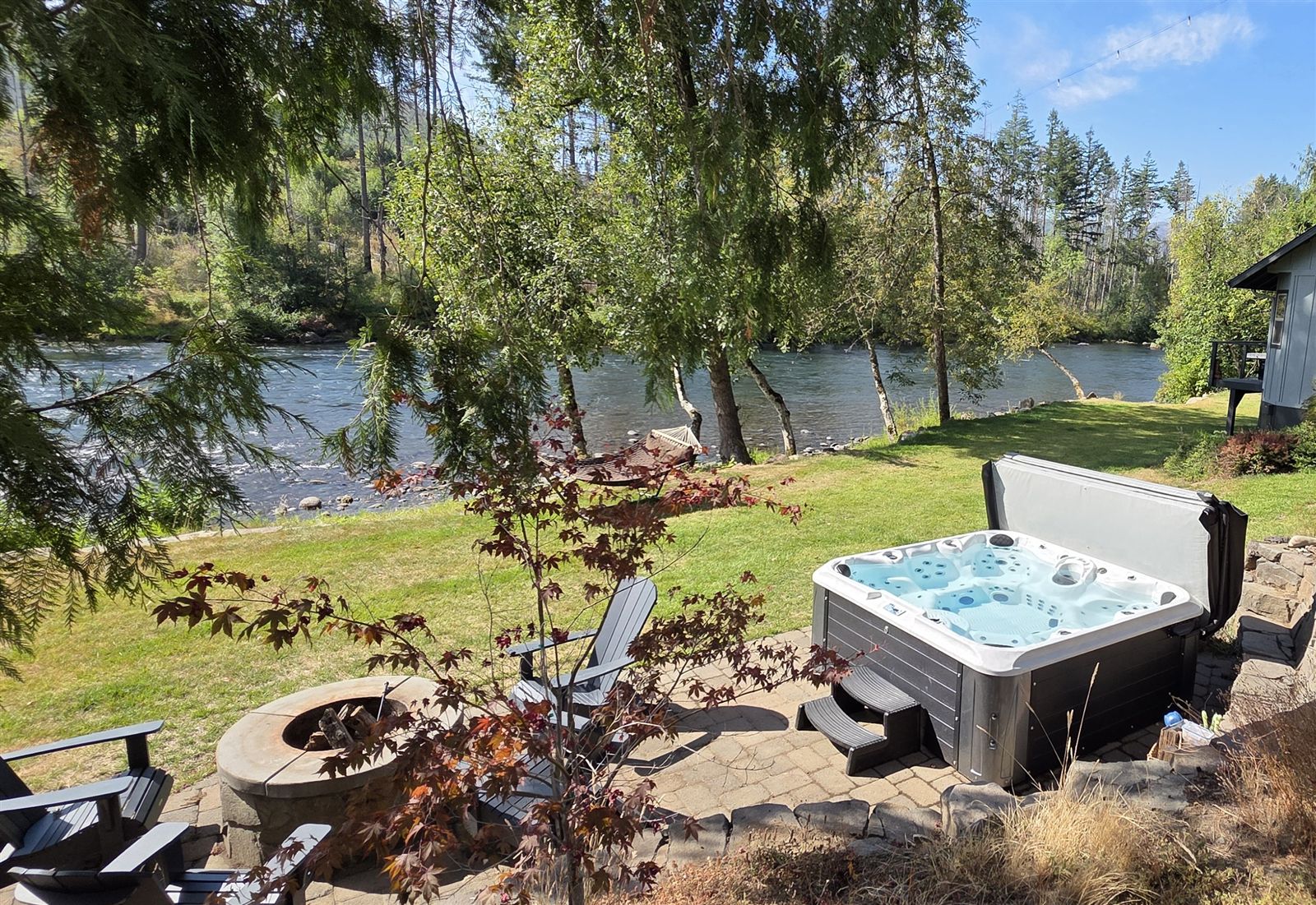 Stone fire pit, Adirondack chairs and hot tub on the lawn beside the McKenzie River at The River Front House